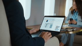 Focused businessman analyzing financial data on a laptop while traveling on a luxurious private jet, with a female colleague sitting across the table using her smartphone during the business trip - Powered by Shutterstock - Get 15% off with code: PIKWIZARD15