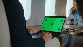 Unrecognizable businessman typing on a laptop with a green screen and tracking markers during a luxurious flight while his female colleague uses a smartphone in the background - Powered by Shutterstock - Get 15% off with code: PIKWIZARD15