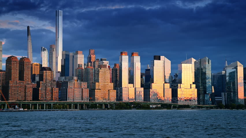 Massive high-rises in the dense skyline of New York reflect the light of setting sun. Dramatic cloudscape over the city. View from the waterscape.