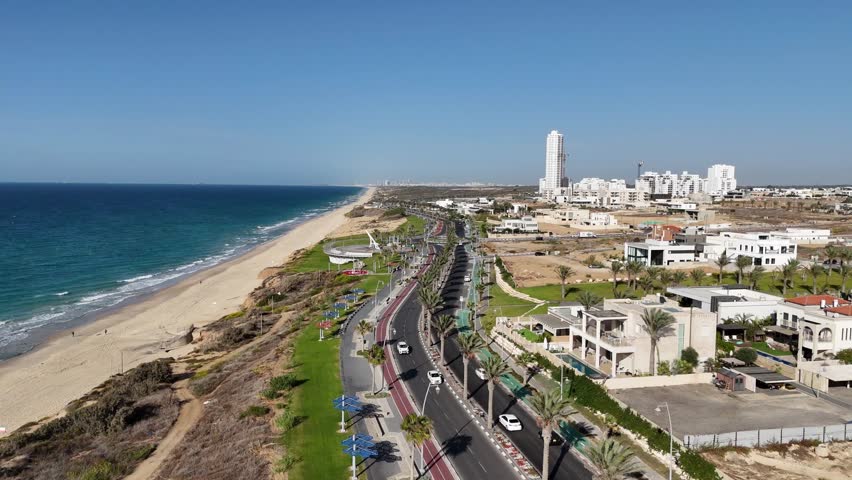 Aerial view of Ashkelon showing busy traffic, residential buildings, and the scenic seaside promenade. Dynamic urban coastline scene with cars in motion and modern city architecture.