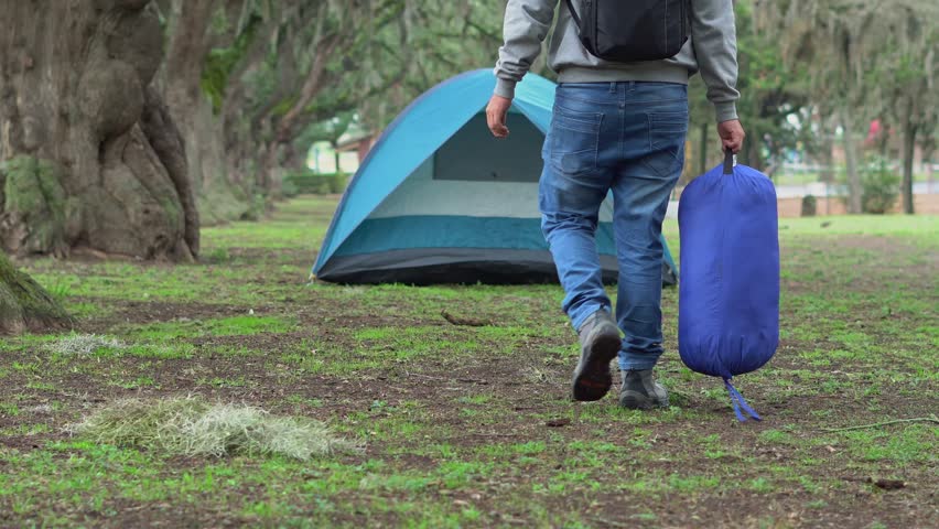 Young man wearing sports clothes and a backpack, carrying a rolled sleeping bag while walking toward his tent in a forest campsite. The scene reflects adventure, outdoor life, and connection with natu