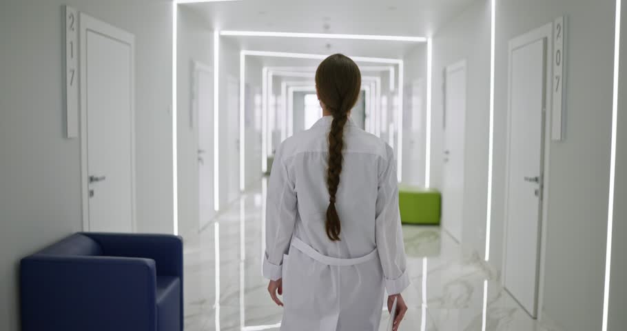Rear view of Female doctor in a white coat with a tablet in her hands walking along the offices in a spacious white corridor in a hospital