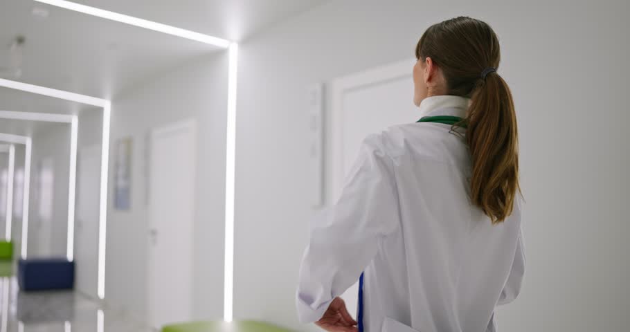Rear view of a female doctor in a white coat walking along the offices in the corridor of a white hospital