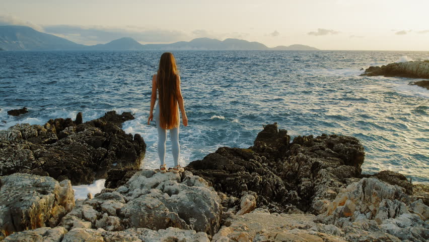 Woman standing on rugged rocks looking at the sea during sunset in Kefalonia