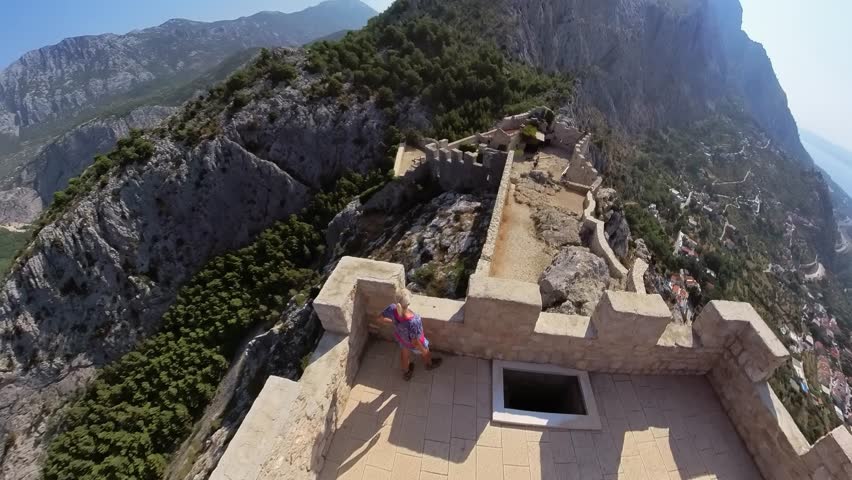 Traveler on Starigrad fortress ramparts overlooking Omis, rugged mountains, dense green hills and the Adriatic coast, panoramic sunny summer view of historic Dalmatian landscape in Croatia