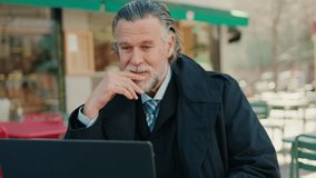 A mature businessman in a suit sits at an outdoor cafe, working on his laptop and having a video conference call. He waves and discusses business. - Powered by Shutterstock - Get 15% off with code: PIKWIZARD15