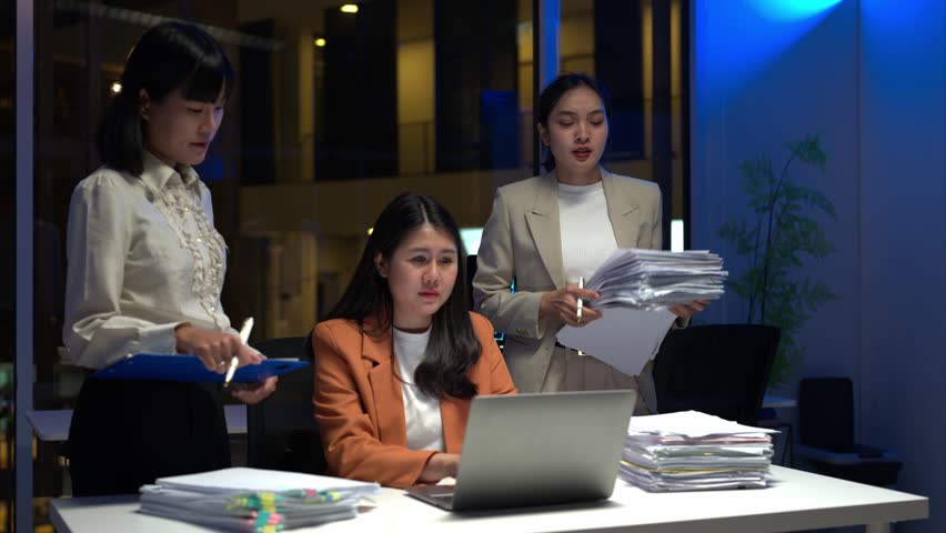 Three women are sitting at a desk with a laptop and piles of papers. They appear to be working together on a project