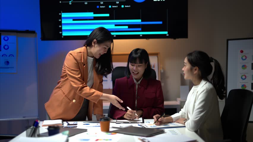 Three women are sitting at a table with a projector screen behind them. One of the women is pointing at something on the screen
