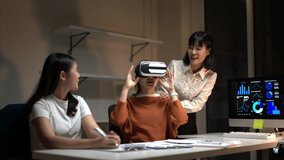 Three women are sitting at a desk with a computer monitor and a virtual reality headset. One of the women is wearing a white shirt and the other two are wearing orange shirts. They are all smiling - Powered by Shutterstock - Get 15% off with code: PIKWIZARD15