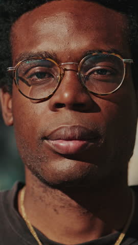 Vertical video, Portrait of a happy young man smiling at the camera outdoors on a sunny day. He is wearing glasses and a gold chain.