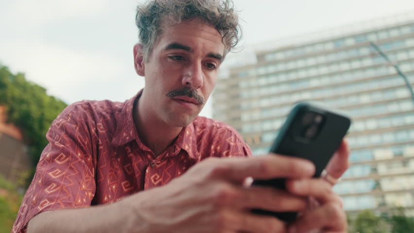 A young man with curly hair and a mustache sits outside and uses his phone. He scrolls, reads, and reacts. City buildings are visible in the background.