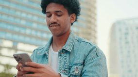 A cheerful young man with curly hair happily interacts with his smartphone while relaxing outdoors. He smiles as he scrolls, enjoying modern connectivity in an urban environment - Powered by Shutterstock - Get 15% off with code: PIKWIZARD15