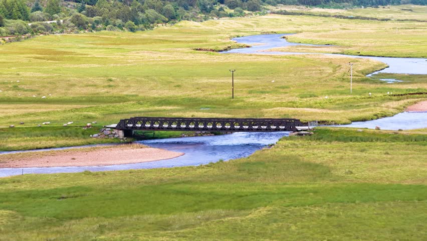 Drone pans across tranquil river, metal bridge, and green valley under soft daylight in Scotland