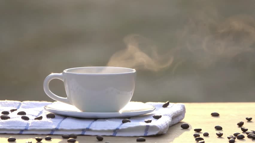Steaming cup of coffee on a table with coffee beans and a towel in soft sunlight