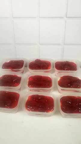 A close-up of a hand wearing a plastic glove holding fresh strawberry slices for topping. A row of no-bake strawberry cheesecakes is in the background.