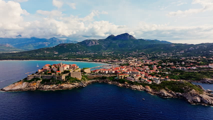 Aerial drone shot over the historic old town of Calvi, Corsica, France. Picturesque view of coastal town, landscape, fluffy clouds, and mountains in the background.