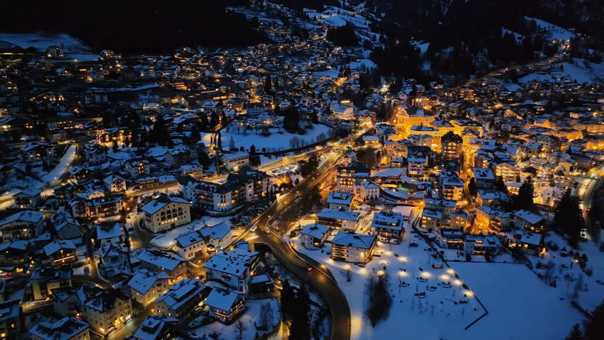 Aerial drone view of Ortisei town in the Dolomites at night, blanketed in snow and glowing with warm lights amid the majestic winter mountain landscape.