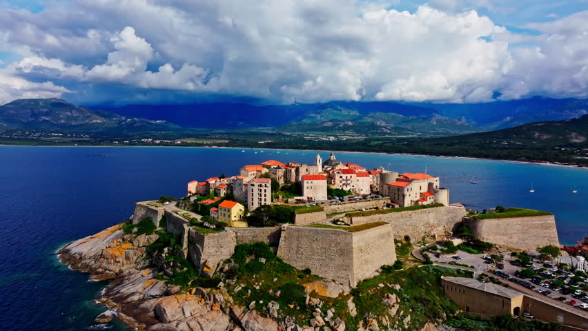 Aerial drone shot over the historic old town of Calvi, Corsica, France. Picturesque view of coastal town, landscape, fluffy clouds, and mountains in the background.