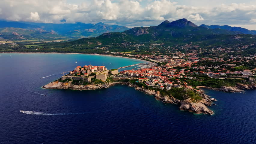 Aerial drone shot over the historic old town of Calvi, Corsica, France. Picturesque view of coastal town, landscape, fluffy clouds, and mountains in the background.