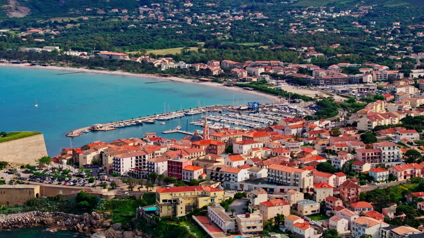 Aerial drone shot over the historic old town of Calvi, Corsica, France. Picturesque view of coastal town, landscape, fluffy clouds, and mountains in the background.
