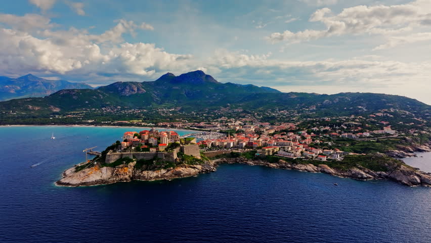Aerial drone shot over the historic old town of Calvi, Corsica, France. Picturesque view of coastal town, landscape, fluffy clouds, and mountains in the background.