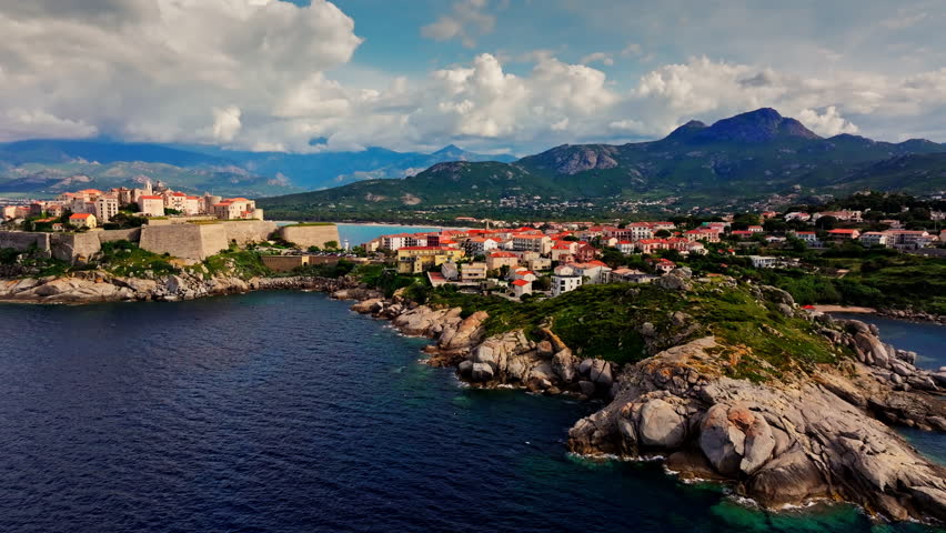 Aerial drone shot over the historic old town of Calvi, Corsica, France. Picturesque view of coastal town, landscape, fluffy clouds, and mountains in the background.
