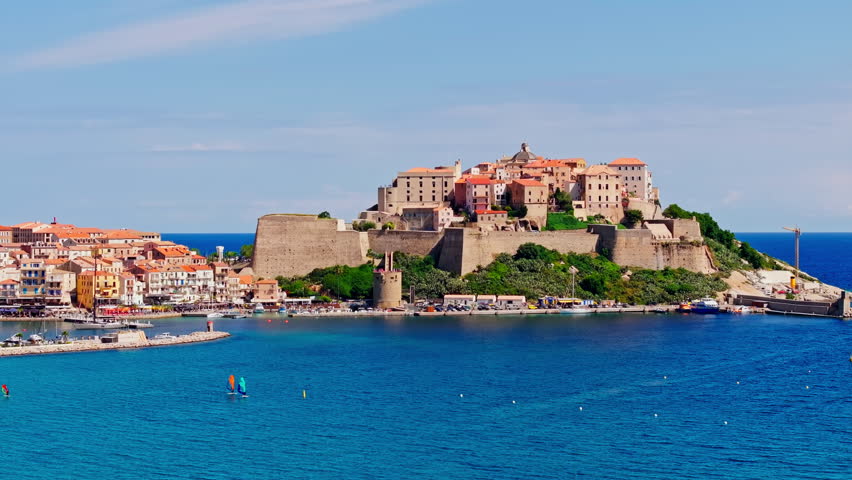 Aerial drone shot over the coastal town of Calvi in Corsica. View of the citadel fortress overlooking the city. Summer holidays destination. Bright blue sky, vibrant turquoise sea.