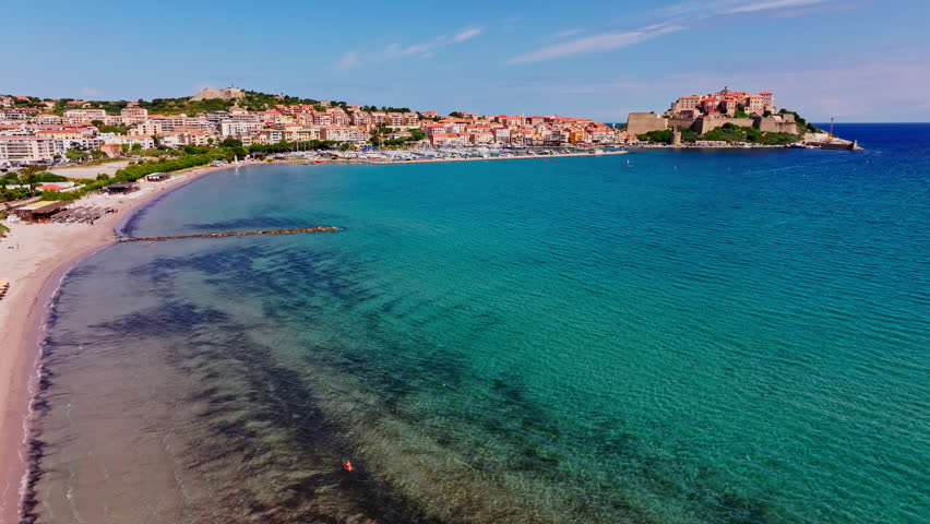 Aerial drone shot over the coastal town of Calvi in Corsica. View of the citadel fortress overlooking the city. Summer holidays destination. Blue sky, vibrant turquoise sea, long stretching beach.