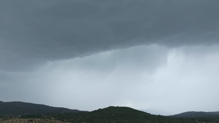 Drone shot panning right across a sky filled with dark storm clouds over shaded hills. Rain descends in distant streaks, bringing life-giving water to the land beneath a dramatic, powerful sky.