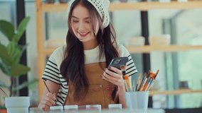 Happy Asian female artist using smartphone to find references and ideas for painting ceramic pottery design in creative studio, woman painting custom artwork for client project - Powered by Shutterstock - Get 15% off with code: PIKWIZARD15