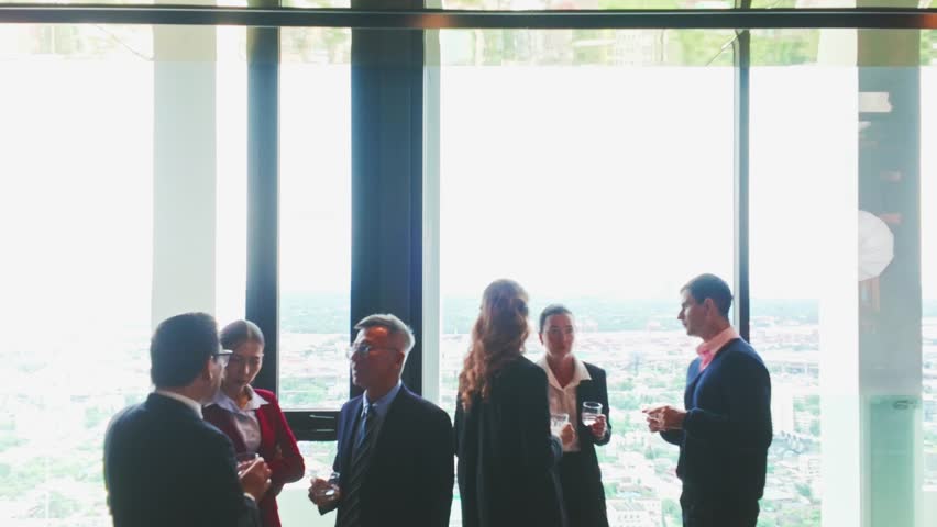 Diverse group of business professionals enjoying conversation during a casual office party or post-meeting gathering. Standing near large windows, they hold drinks and chat in a relaxed mood.