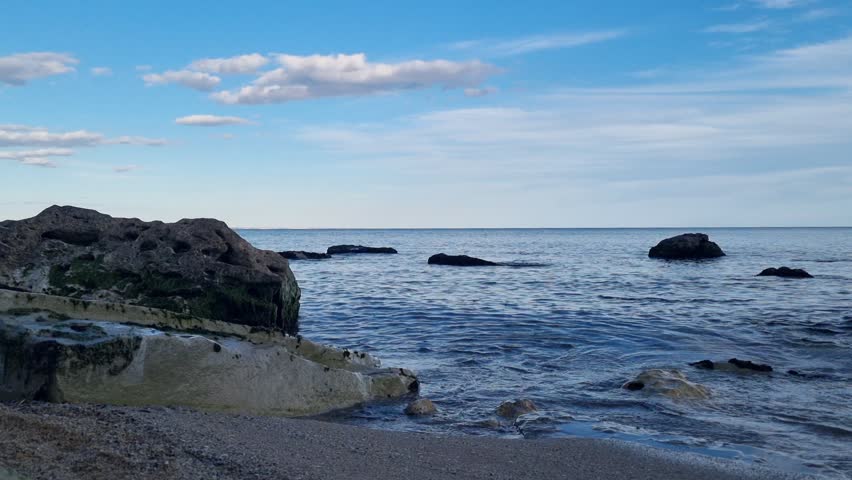 Beautiful seashore view with gentle waves washing over rocks under a bright blue sky. Peaceful ocean landscape perfect for relaxation, travel videos, meditation, and nature backgrounds.