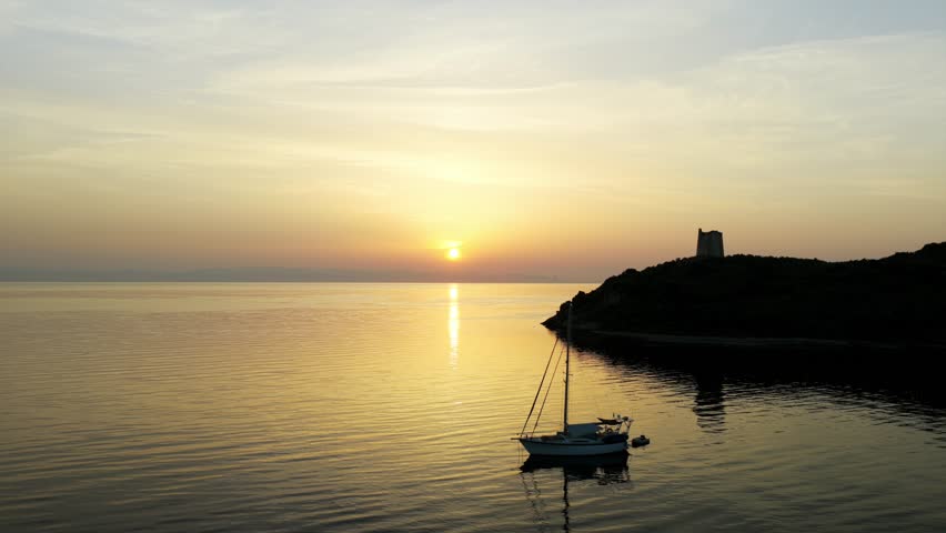 Sailboat at anchor in calm bay at sunset, coastal tower silhouetted on headland, Sardinia, Italy. Aerial drone lateral view