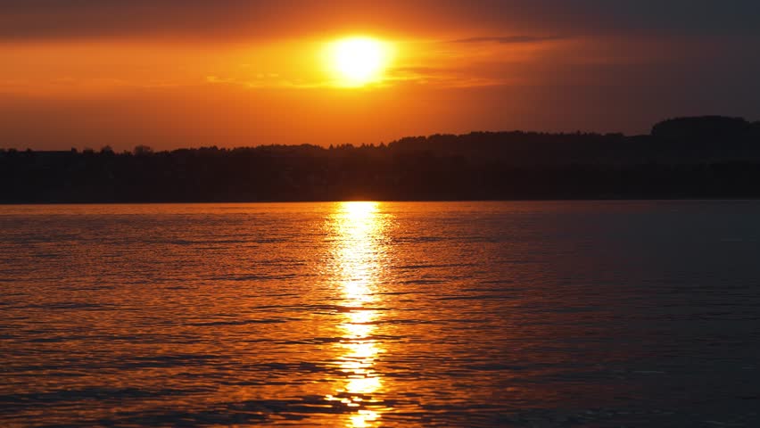 Beautiful orange sun setting over the dark horizon, casting a shimmering golden reflection on the calm water surface of a lake, creating a peaceful and romantic evening atmosphere