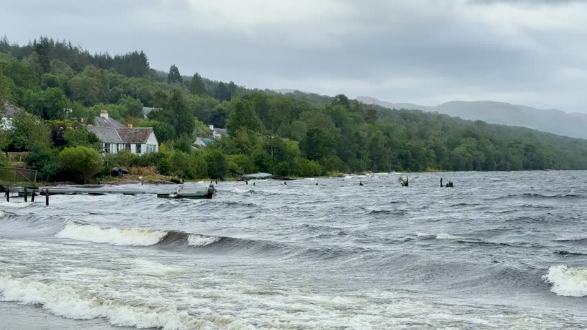 Choppy waves crash on Loch Ness shore, cloudy skies, green hills, static wide shot, daytime