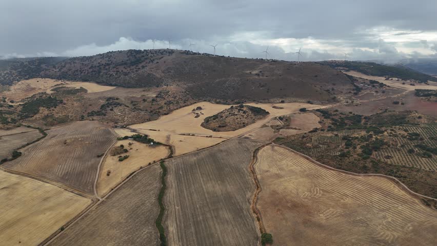 Aerial view of the landscape around Turón Castle in Ardales, Málaga, Spain, featuring dry farmland, scattered trees, and wind turbines under a cloudy sky.