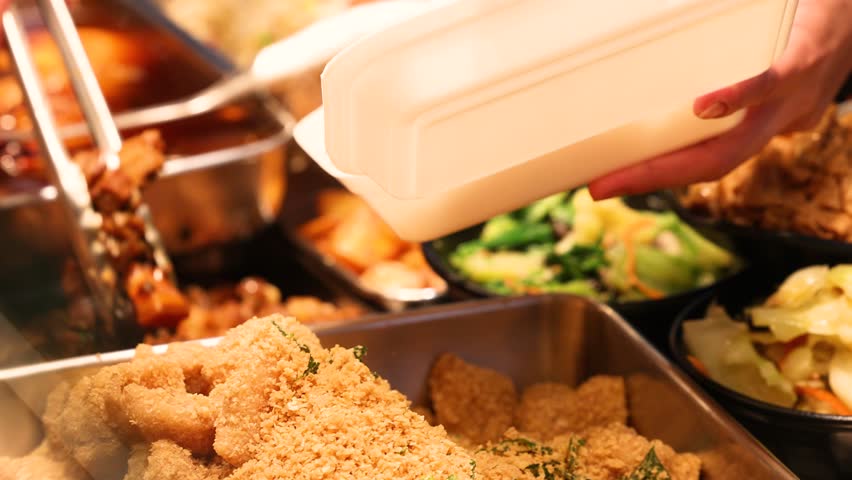 Close-up of tongs picking various Asian vegetable and meat dishes at a food court, placing them into disposable takeout containers, illustrating custom meal selection and convenient urban dining