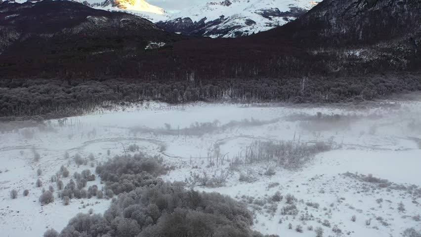 Aerial side shot moving from right to left showing the snowy valley of Haruwen in Tierra del Fuego, Argentina, with frozen trees, mist, and mountains in the background lit by sunlight.