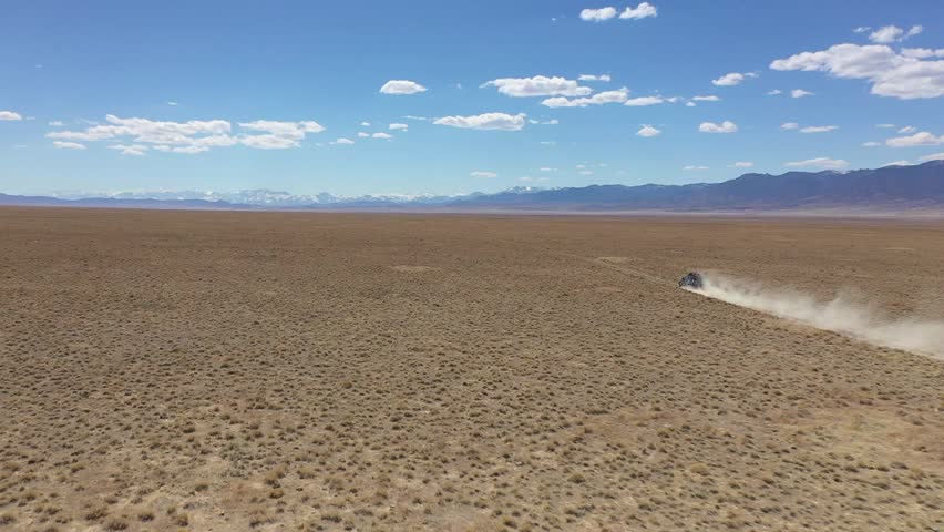 Aerial drone shot following an off-road vehicle driving along a dusty track through the arid Pony Express Valley in Nevada, with distant snowy mountain peaks on the horizon.