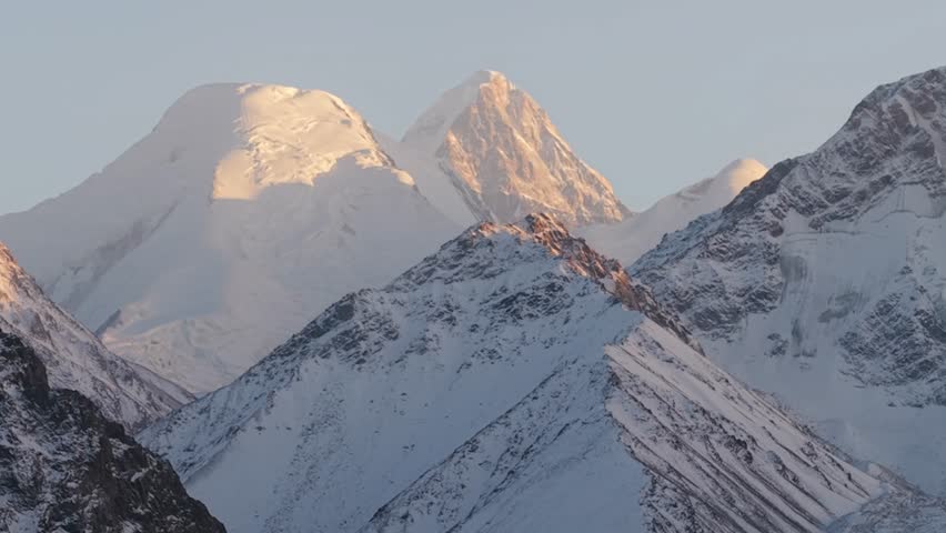 Majestic snow-capped mountains dominate the scene as the camera pans right, showcasing the intricate textures of rocky slopes and glistening snow under soft sunlight, revealing breathtaking peaks