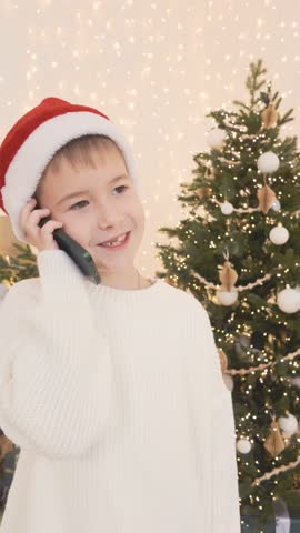 Happy seven year old boy in Santa hat talking on phone call near Christmas tree at home