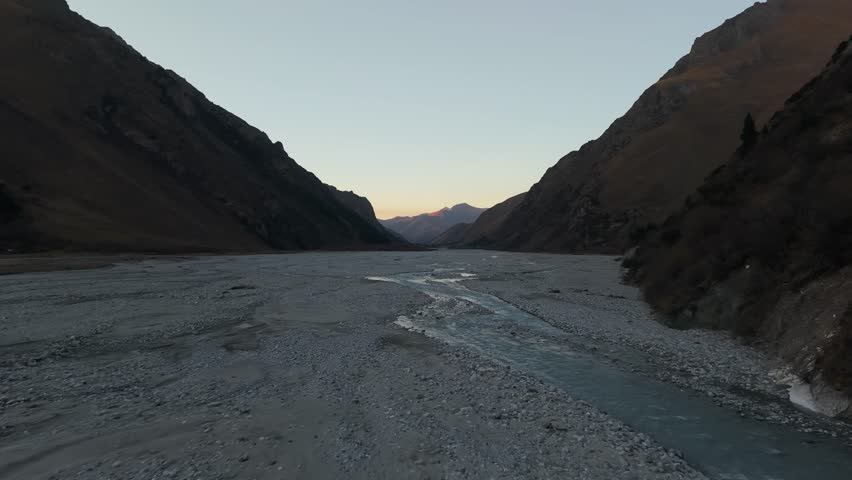 River meanders through a rocky valley as dusk settles, showcasing the rugged terrain and soft evening light, the camera pans smoothly to capture the expansive scenery and tranquil atmosphere