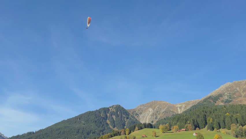 One paraglider soaring through a clear blue sky over a scenic mountain valley in the alps