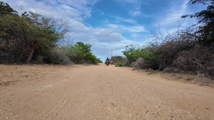 Low-angle, static shot of a fully equipped dual-sport motorcyclist riding directly toward the camera on a dusty, rugged path in a dry, scrubland environment.