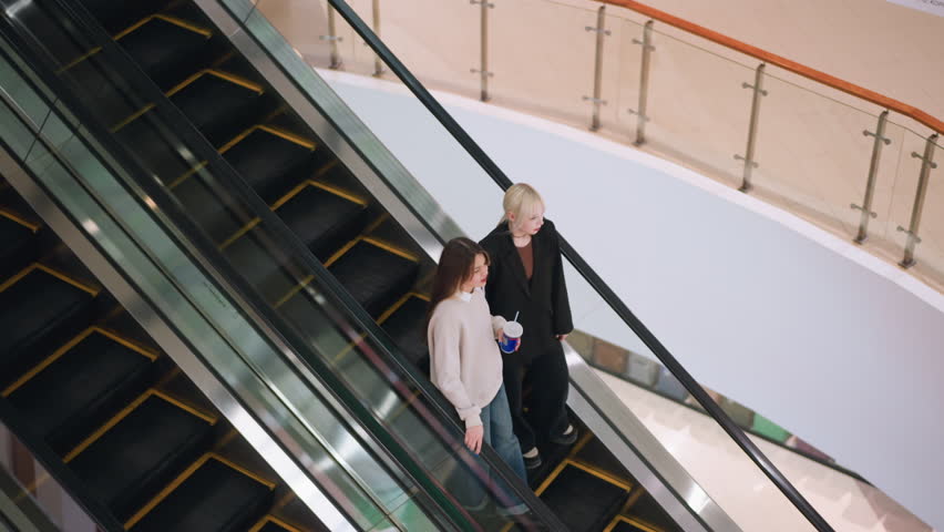 Aerial view of two ladies descending on escalator inside shopping mall, engaging in casual conversation while one holds drink, creating relaxed lifestyle atmosphere in modern indoor environment