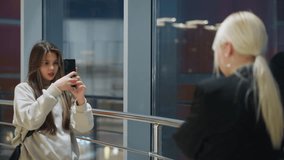 Young lady holding phone carefully taking photo of her sister indoors near large glass window while colorful city lights blink and reflect on glass inside the mall - Powered by Shutterstock - Get 15% off with code: PIKWIZARD15