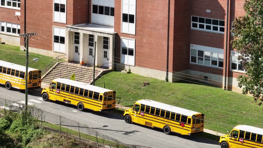 Pick up Line of yellow school buses in front of red brick school on american. Sunny day in fall season. Aerial zoom shot.