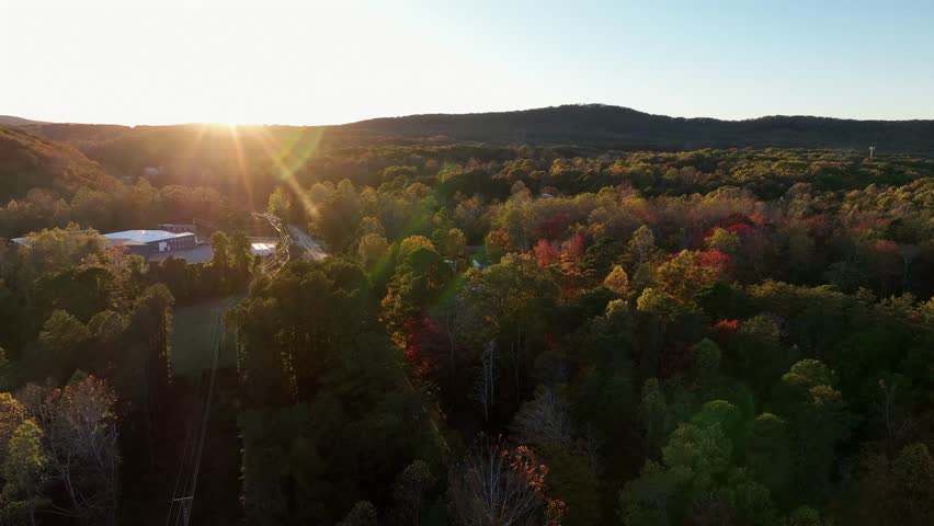 Aerial flyover company in rural suburb district of american town. Golden sunset time lighting over multi colored trees in fall season. Wide shot. Sunset behind mountains in USA.