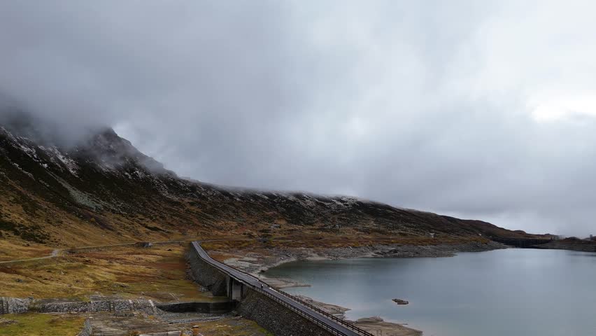 A breathtaking aerial droen footage of a narrow road winding through a serene lake in the heart of the Alps at Splügen Pass.Surrounded by towering mountains and crystal-clear water.