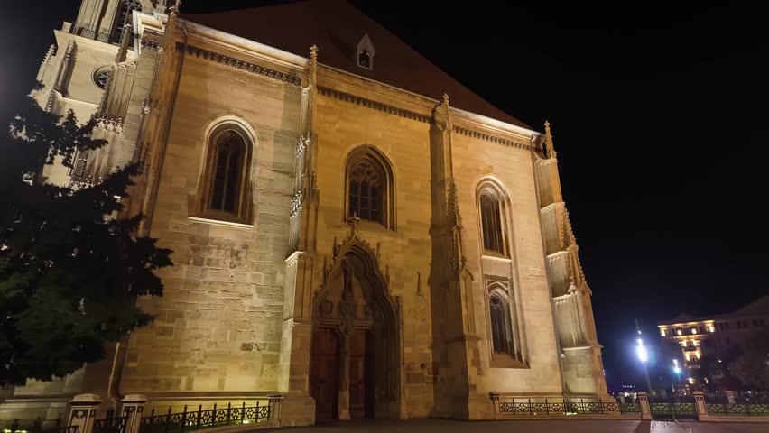 Nighttime view of the illuminated Gothic architecture of St. Michael’s Church in Cluj-Napoca, Romania, captured from the side while walking around the historic landmark in Union Square.